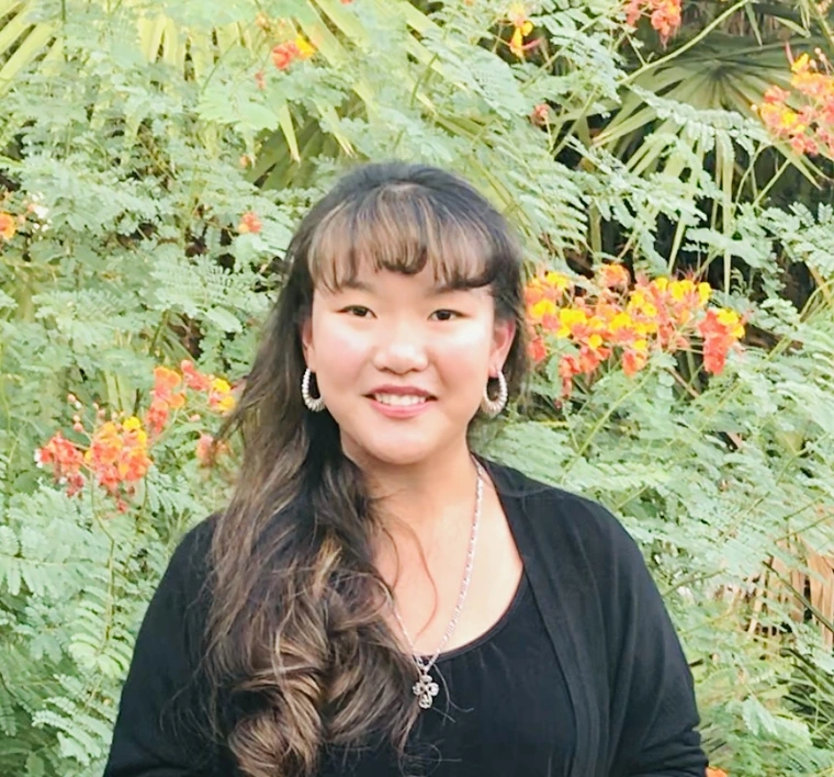A person with long dark hair standing against a green foliage background
