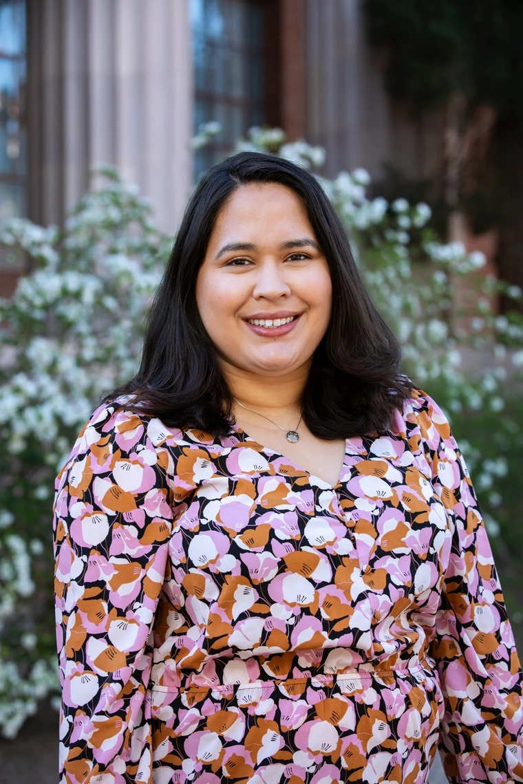 A person with shoulder length dark hair stands against a floral background.